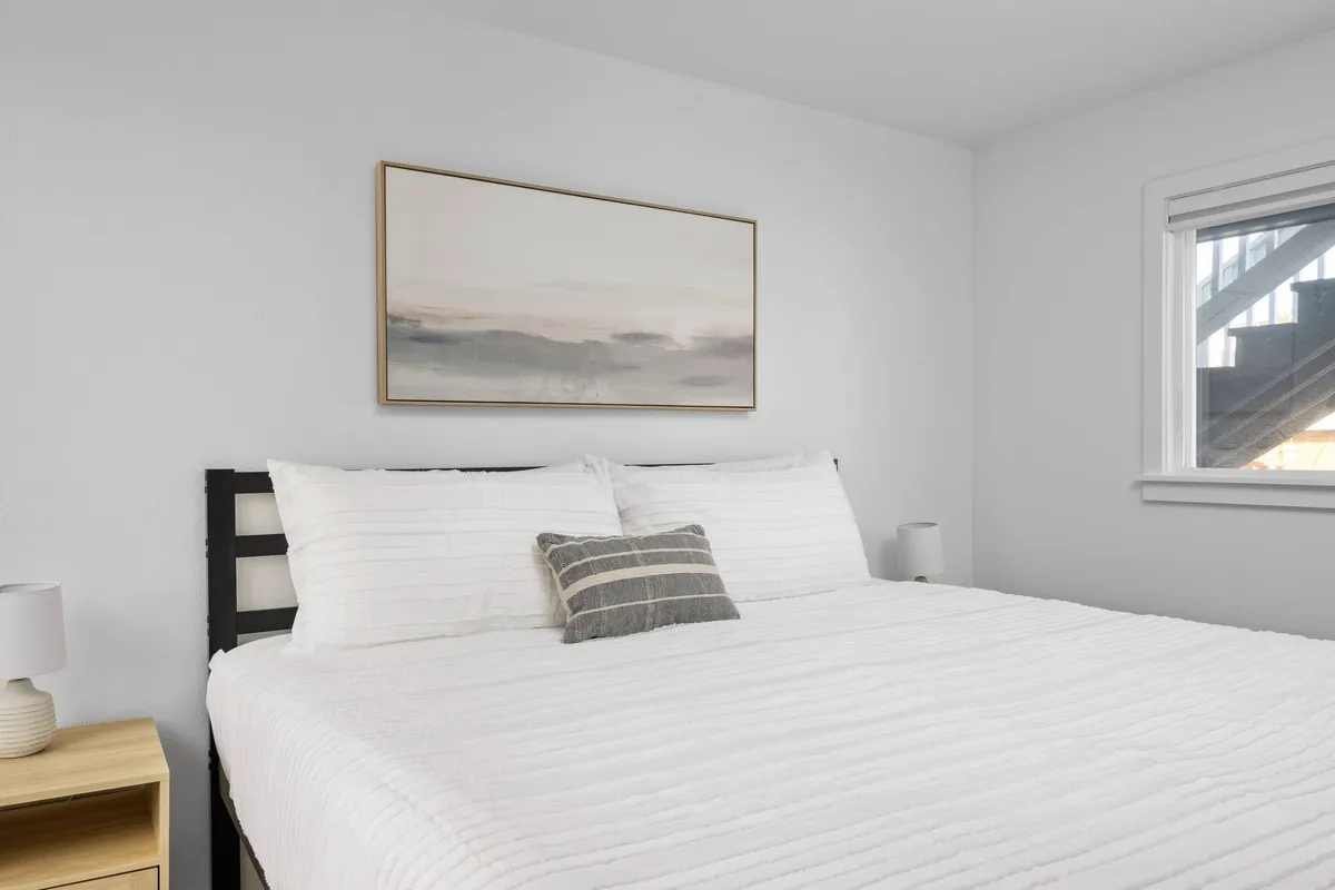 A cozy, inviting sleeping area with a well-made bed, elegant wall art, and a softly glowing nightstand lamp. Gentle natural light streams across the room, highlighting the clean, peaceful design and thoughtful touches that invite relaxation and restful sleep.