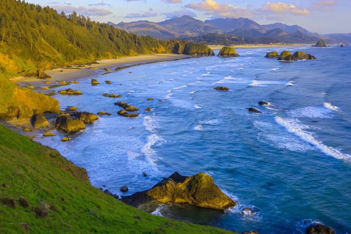 A breathtaking view of the rocky coastline with gentle waves, distant mountains, and the sky bathed in golden sunset hues. The scene is tranquil and expansive, ideal for capturing the mood of a peaceful beachside retreat. Activities like photography, wildlife watching, and quiet contemplation are suggested by the serene setting.