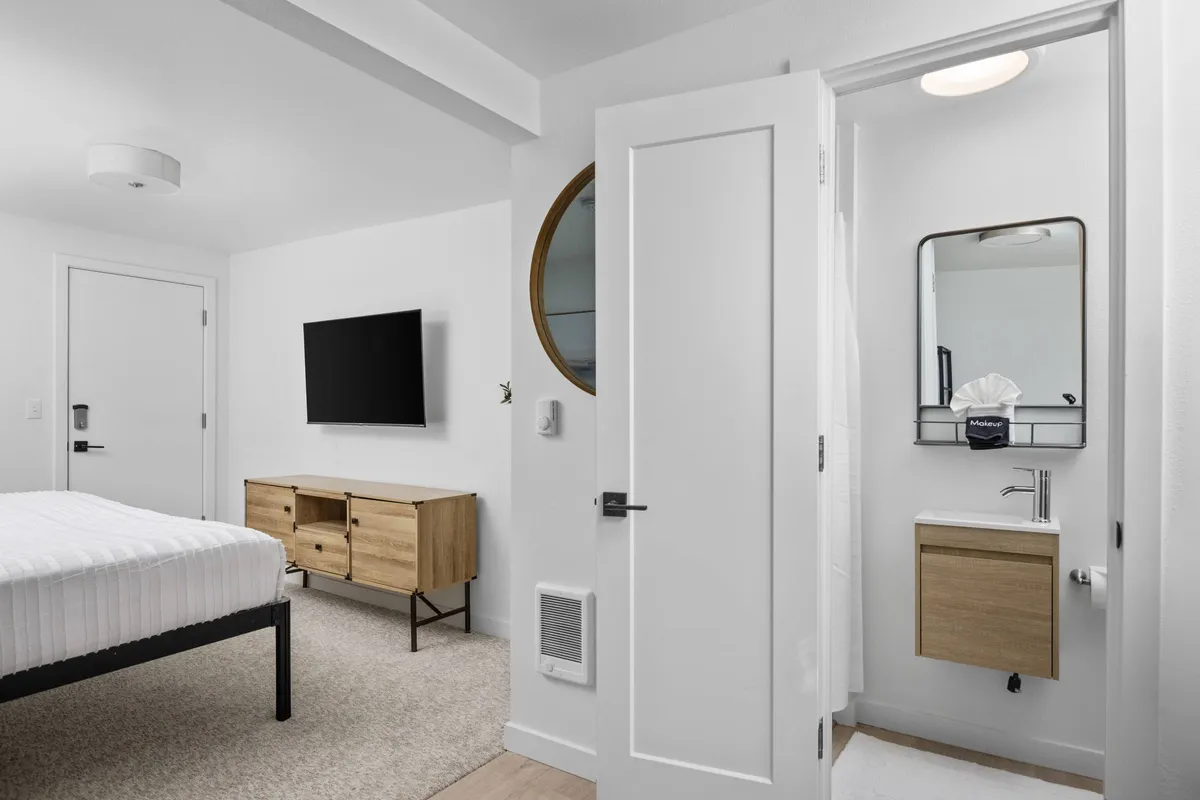 A serene, well-appointed bedroom featuring a neatly made bed, a TV mounted on the wall, a bathroom door slightly ajar, a mirror reflecting natural light, and modern wooden furniture. The space evokes a sense of order and comfort, with contemporary finishes that highlight recent remodeling.