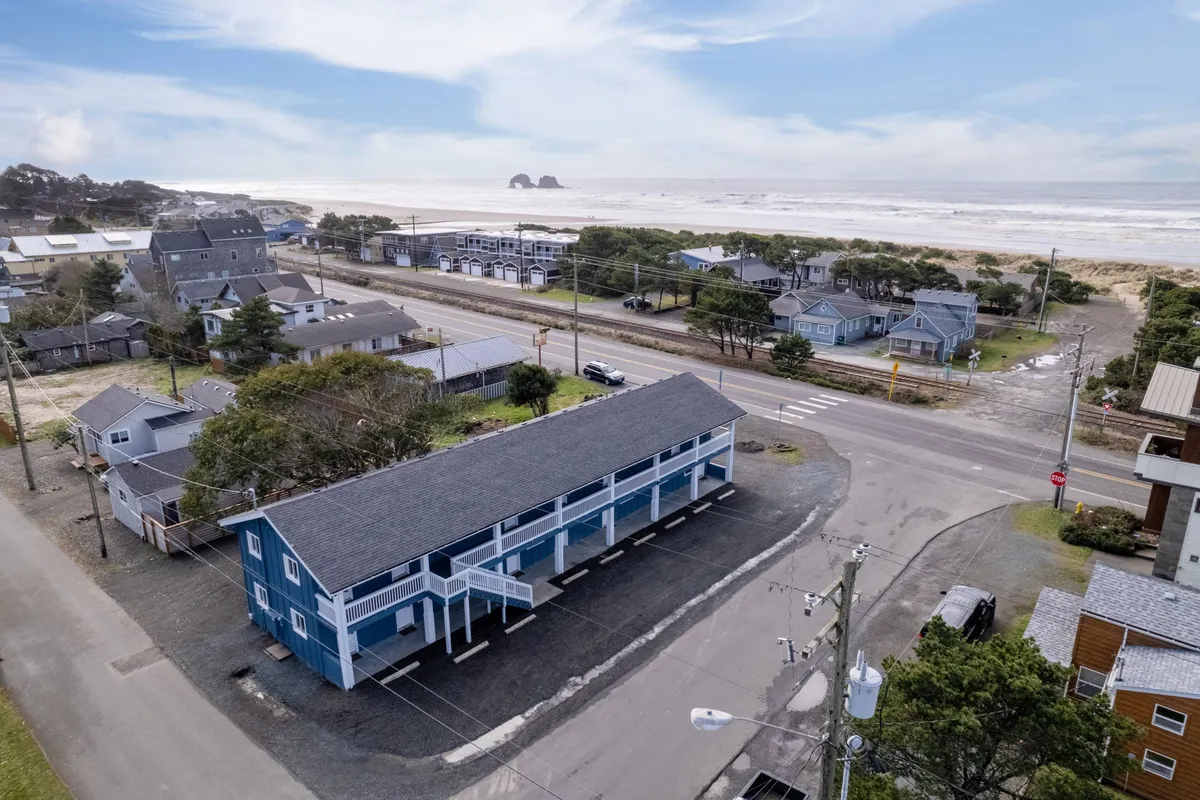 This image shows a relaxed, welcoming view of a blue building with the ocean in the background. The scene includes surrounding houses and a roadway leading toward the shore. People can be seen enjoying beach activities like beachcombing, strolling along the sand, and watching the sunset, giving a sense of easy access to the beach right from your doorstep.