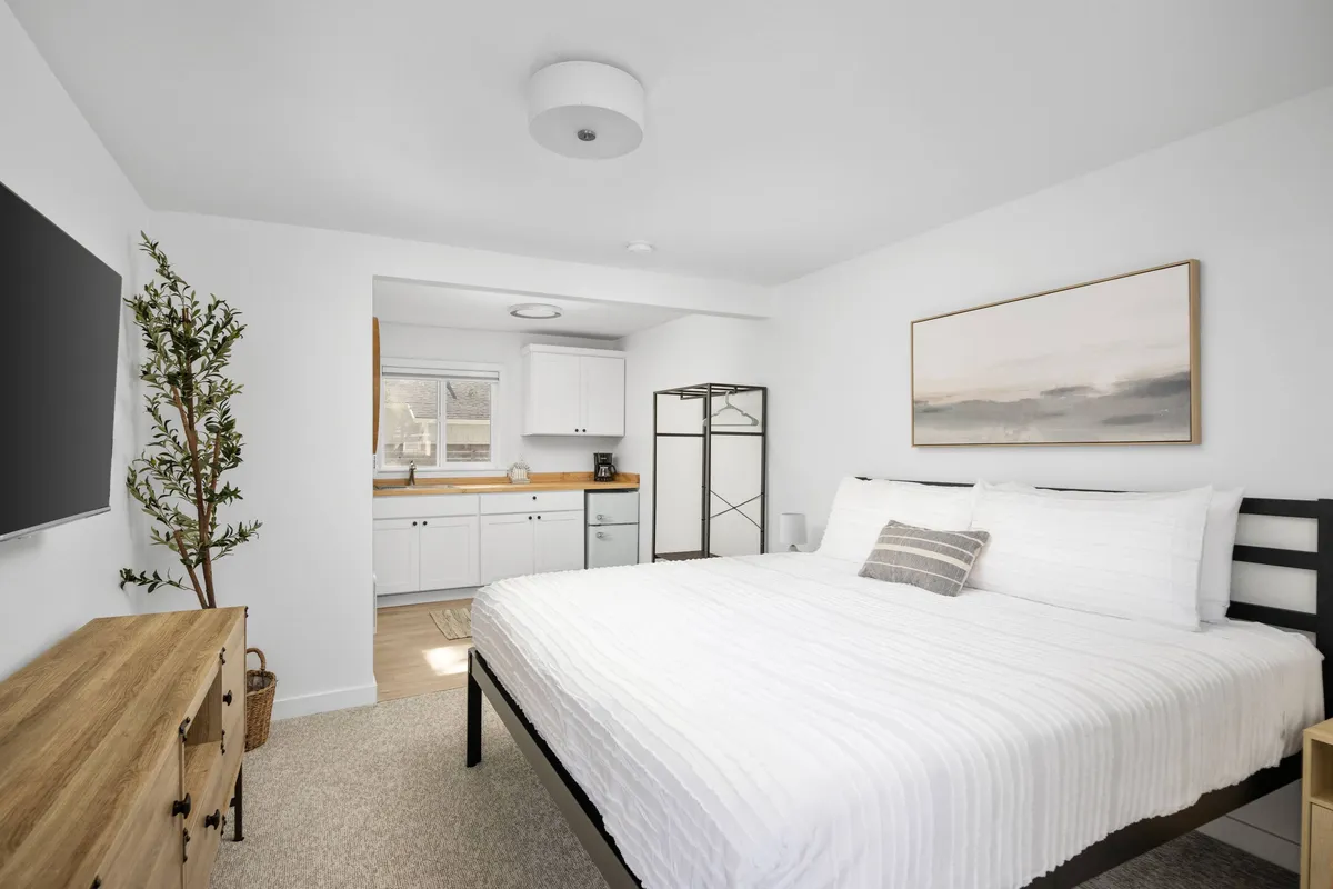 The hero image showcases a serene, light-filled bedroom with a large, inviting bed, a minimalist dresser, a vibrant green plant adding a touch of nature, and a compact kitchenette ideal for simple meals. The space feels modern, uncluttered, and relaxing, with soft natural light streaming in to create a peaceful atmosphere.