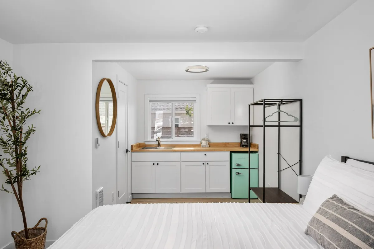 This image showcases a serene interior scene featuring a bed with crisp white linens positioned near a compact kitchenette area. The kitchenette includes a green cabinet and a round mirror, suggesting a stylish yet functional design. Morning sunlight streams in, highlighting the inviting and multi-purpose nature of the space where both relaxation and meal preparation are possible.